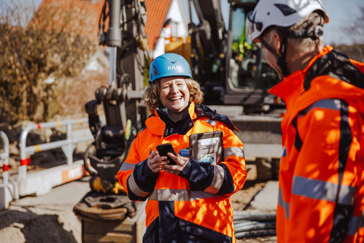 Två projektledare i hjälm och arbetskläder skrattar vid ett ledningsarbete på en gata. 
