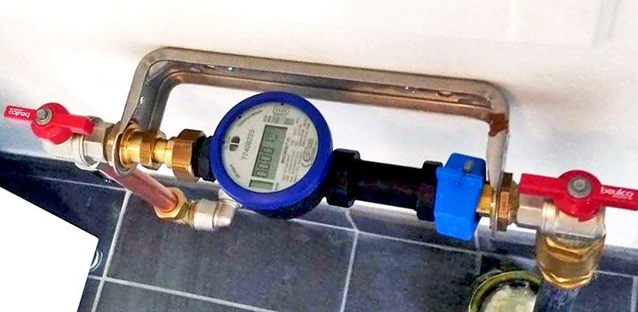 Vattenmätarplats inomhus i uppvärmt utrymme, bestående av vattenmätare, ledningar, ventiler och väggkonsol.